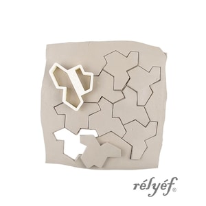 Peut inclure: Un bloc d'argile gris clair avec un motif de puzzle géométrique. Une pièce de puzzle blanche est placée sur l'argile, avec plusieurs pièces de puzzle découpées. Le mot "rélyéf" est imprimé en écriture cursive.