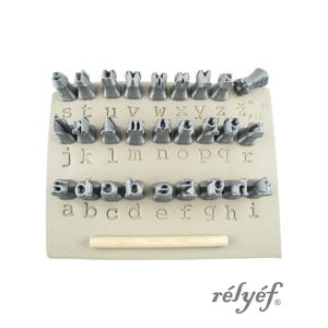 May include: A set of gray metal letter stamps arranged on a beige rectangular base, with a wooden handle at the bottom. The letters are in both uppercase and lowercase. The brand name "rélyéf" is printed in gray.