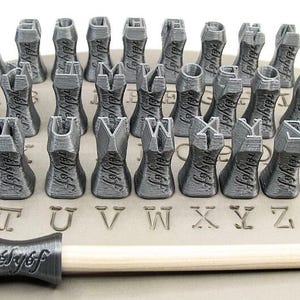 K&ouml;nnte beinhalten: Ein Satz von 26 grauen Alphabet-Stempeln mit einem Holzgriff. Die Stempel sind alphabetisch geordnet und dienen zum Erstellen von Abdr&uuml;cken in Ton oder anderen weichen Materialien. Die Stempel sind seitlich mit "relyef" beschriftet.