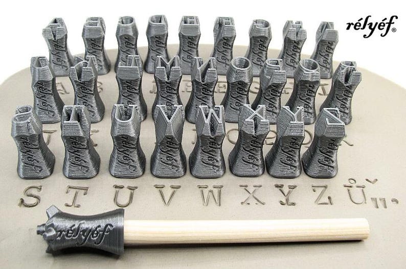 K&ouml;nnte beinhalten: Ein Satz von 26 grauen Alphabet-Stempeln mit einem Holzgriff. Die Stempel sind alphabetisch geordnet und dienen zum Erstellen von Abdr&uuml;cken in Ton oder anderen weichen Materialien. Die Stempel sind seitlich mit "relyef" beschriftet.