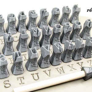 K&ouml;nnte beinhalten: Ein Satz von 26 silberfarbenen Metallstempeln in Form von Buchstaben des Alphabets, die in alphabetischer Reihenfolge angeordnet sind, mit dem Wort "r&eacute;ly&eacute;f" auf einer wei&szlig;en Oberfl&auml;che.