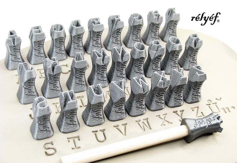 K&ouml;nnte beinhalten: Ein Satz von 26 silberfarbenen Metallstempeln in Form von Buchstaben des Alphabets, die in alphabetischer Reihenfolge angeordnet sind, mit dem Wort "r&eacute;ly&eacute;f" auf einer wei&szlig;en Oberfl&auml;che.