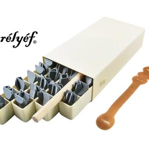 May include: A set of 25 metal letter stamps in a beige box with a wooden handle. The box has the word "r&eacute;ly&eacute;f" printed on the front. The stamps are arranged in a grid pattern and are used for stamping letters onto various surfaces.
