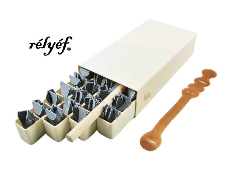 May include: A set of 25 metal letter stamps in a beige box with a wooden handle. The box has the word "r&eacute;ly&eacute;f" printed on the front. The stamps are arranged in a grid pattern and are used for stamping letters onto various surfaces.