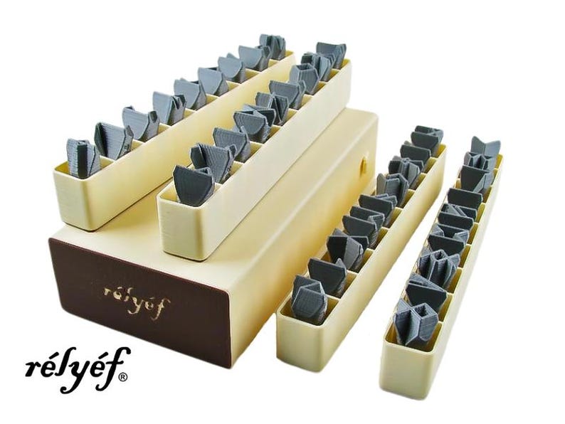 May include: A set of 36 gray plastic embossing stamps organized in three tiers of beige plastic trays. The trays are labeled "r&eacute;ly&eacute;f" in brown lettering.