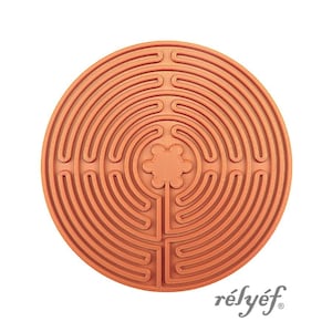 Può includere: Un oggetto circolare color terracotta caratterizzato da un motivo a labirinto in rilievo con una forma di fiore a sei petali al centro. Il design è composto da percorsi concentrici che portano al centro. Il testo "rélyéf" è stampato nell'angolo in basso a destra.