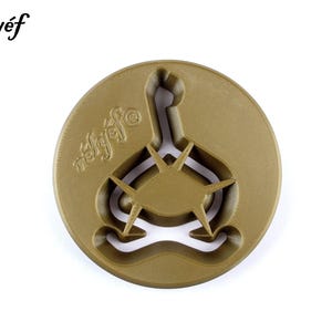 May include: A gold-colored, circular cookie cutter with a unique design. The cutter features a stylized shape with a central oval and radiating points, and the word "rélyéf" is printed on the top left. The design is suitable for baking.