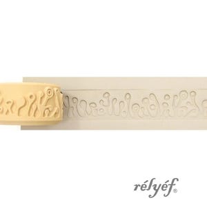 Puede incluir: Un sello en relieve beige con un diseño abstracto y la palabra "rélyéf" en escritura gris. El sello tiene un patrón repetitivo de formas orgánicas. El diseño está tallado en un bloque rectangular.