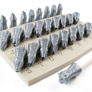 May include: A set of 26 silver-colored metal alphabet stamps with a wooden handle. The stamps are arranged on a beige clay surface with the letters of the alphabet printed on it. The stamps are used for stamping letters into clay or other materials.