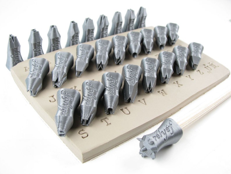 May include: A set of 26 silver-colored metal alphabet stamps with a wooden handle. The stamps are arranged on a beige clay surface with the letters of the alphabet printed on it. The stamps are used for stamping letters into clay or other materials.
