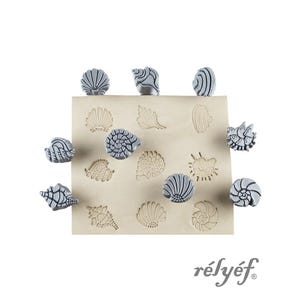May include: A collection of gray seashell-shaped stamps and imprints on a beige surface. The stamps are arranged around the edges of the surface, with corresponding imprints in the center. The word "rélyéf" is printed in a cursive font.