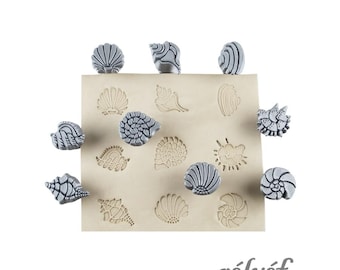 May include: A collection of gray seashell-shaped stamps and imprints on a beige surface. The stamps are arranged around the edges of the surface, with corresponding imprints in the center. The word "rélyéf" is printed in a cursive font.