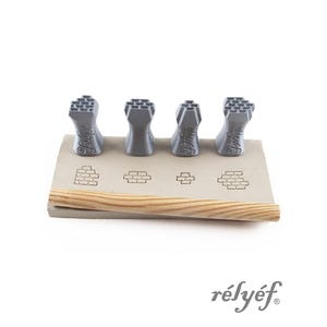 Könnte beinhalten: Vier graue, 3D-gedruckte, ziegelsteinförmige Stempel sind auf einer rechteckigen, hellbraunen Oberfläche mit Ziegelabdrucken angeordnet. Unten befindet sich ein Holzdübel. Der Markenname "rélyéf" ist grau gedruckt.
