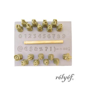 May include: A set of gold-colored metal stamps with numbers, letters, and symbols. The stamps are arranged on a gray rectangular base. The numbers 0-9 are included, along with various punctuation marks and symbols.