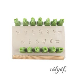 May include: A set of green rubber stamps with various botanical designs, including flowers, leaves, and a small pot. The stamps are arranged on a beige surface, with a wooden handle at the bottom. The word "rélyéf" is printed in a cursive font.