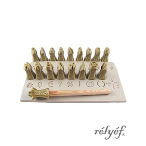 May include: A set of relief stamps with various symbols and designs, including letters, numbers, and punctuation marks. The stamps are arranged on a rectangular base, with a wooden handled mallet. The brand name "rélyéf" is visible.