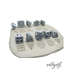 May include: A collection of gray, Celtic knot-design stamps and impressions. The stamps are in various shapes, including squares, triangles, and rounded forms. The impressions are pressed into a light tan clay-like surface. The word "rélyéf" is in the bottom right corner.