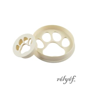 May include: Two off-white paw print-shaped cookie cutters. One is a small, circular cutter, and the other is a larger, round cutter. The cutters are made of a light-colored plastic and are designed for baking. The brand name "rélyéf" is printed in a script font.