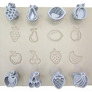Puede incluir: Un conjunto de sellos de arcilla con forma de frutas, que incluyen uvas, peras, limones, melocotones, frambuesas, manzanas, cerezas, fresas y plátanos. Los sellos son grises y están dispuestos sobre una superficie rectangular beige.