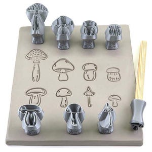 May include: A set of seven grey mushroom-shaped clay cutters and one wooden handle tool arranged on a flat grey clay slab. The clay slab features eight distinct mushroom impressions stamped into the surface, showcasing various cap and stem designs.