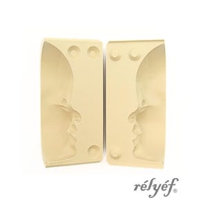 Puede incluir: Dos moldes de silicona beige, cada uno con un perfil de rostro. Los moldes tienen forma rectangular con bordes redondeados y tres hendiduras circulares. La marca "rélyéf" está impresa en cursiva.