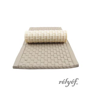 May include: A beige yoga mat with a woven pattern, with a matching cylindrical roller on top. The mat and roller are made of a soft, textured material. The brand name "rélyéf" is printed in a cursive font.