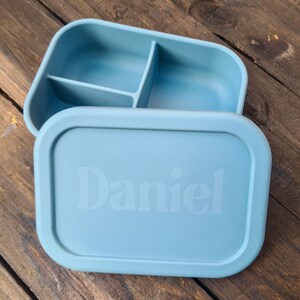 Puede incluir: Una fiambrera de silicona azul claro con tres compartimentos y una tapa. La tapa tiene el nombre "Daniel" grabado en relieve.