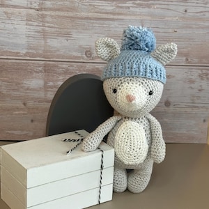 May include: A gray and white crocheted stuffed animal wearing a light blue crocheted hat with a pom-pom. The animal is standing next to a stack of white boxes with black and white string.