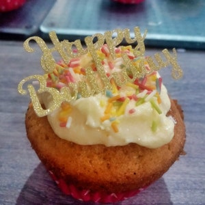 Puede incluir: Un cupcake con glaseado blanco y chispas de colores, coronado con un letrero dorado brillante de "Happy Birthday". El cupcake está en un molde de lunares rojos y blancos. Otros cupcakes son visibles en el fondo.