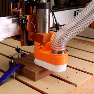 May include: A CNC machine in operation, featuring an orange and silver milling head with a white brush attachment. A wooden block is clamped in place, with a flexible hose extending from the machine. The machine is mounted on a wooden surface.