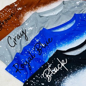 May include: Several t-shirts in shades of brown, grey, royal blue, and black. Each top features a bleached or splattered pattern with white spots. The colour names are written in cursive on each shirt.