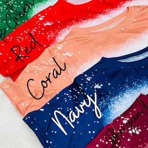 May include: A selection of colourful t-shirts in various shades, including dark green, red, coral, navy, and burgundy. Each shirt features a unique white speckled design. The names of the colours are written in cursive on the shirts.