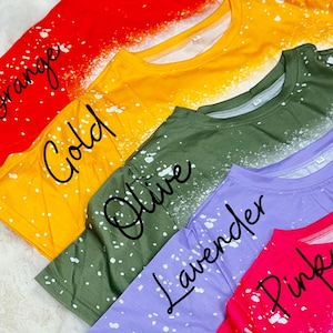 May include: A selection of crew neck t-shirts in orange, gold, olive, lavender, and pink. Each top features a white splatter effect. The colour names are written on the shirts.