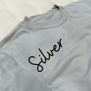 May include: A light silver-gray t-shirt with the word "Silver" in black script. The shirt has a crew neck and short sleeves. The fabric appears soft and slightly wrinkled.