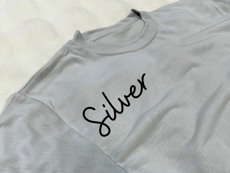 May include: A light silver-gray t-shirt with the word "Silver" in black script. The shirt has a crew neck and short sleeves. The fabric appears soft and slightly wrinkled.