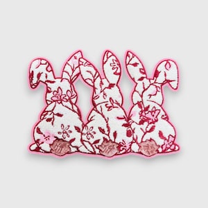 May include: Three white bunny patches with a floral pattern in red and pink, outlined in pink. The bunnies' bottoms have a sequined, pink, circular design. The patches are likely for clothing or craft projects.