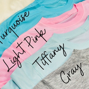 May include: A close-up of four t-shirts in turquoise, light pink, Tiffany blue, and gray. The names of the colors are written in cursive on each shirt. The shirts are folded, showing the neckline.