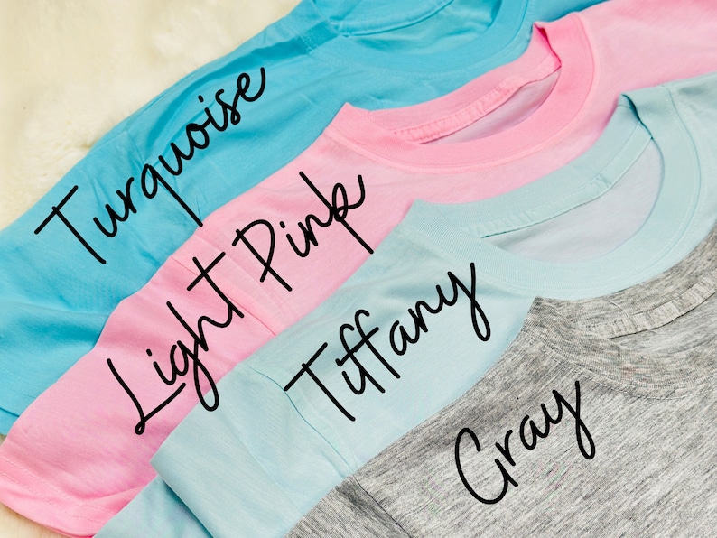 May include: A close-up of four t-shirts in turquoise, light pink, Tiffany blue, and gray. The names of the colors are written in cursive on each shirt. The shirts are folded, showing the neckline.