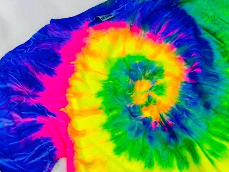May include: A tie-dye shirt with a bright yellow, green, blue, and pink spiral pattern. The shirt has a tag that says "Gildan" on it.