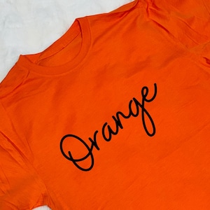 May include: Bright orange t-shirt with the word "Orange" in a black cursive font. The shirt is made of a soft material and has a crew neck. The image is taken from a slightly elevated angle.