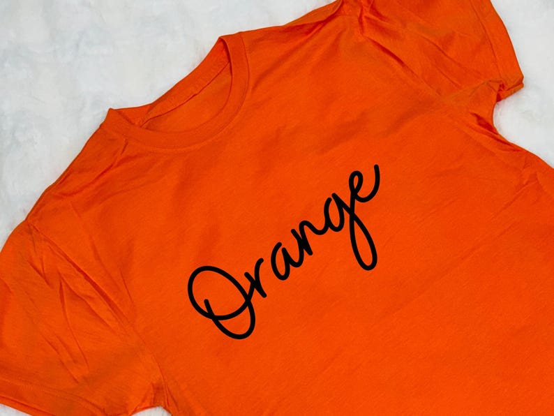 May include: Bright orange t-shirt with the word "Orange" in a black cursive font. The shirt is made of a soft material and has a crew neck. The image is taken from a slightly elevated angle.