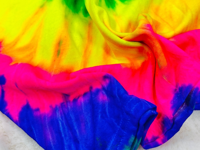 May include: A tie-dye fabric with a bright yellow, orange, pink, blue and green color scheme. The fabric is folded and draped, showing the different colors.