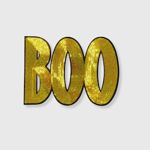 May include: A gold sequined "BOO" sign with a black outline. The letters are filled with small, shiny gold sequins, creating a textured and eye-catching effect. The sign is perfect for Halloween decorations.