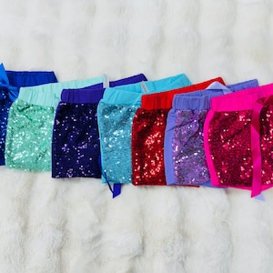 May include: Seven pairs of sequined shorts in various colors, including blue, green, purple, red, and pink. Each pair has a matching ribbon tie at the waist.