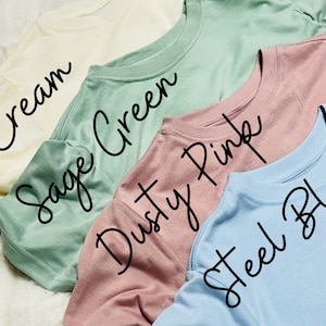 May include: A collection of five crew neck t-shirts in various pastel colors: cream, sage green, dusty pink, and steel blue. The names of the colors are written in cursive across the shirts.