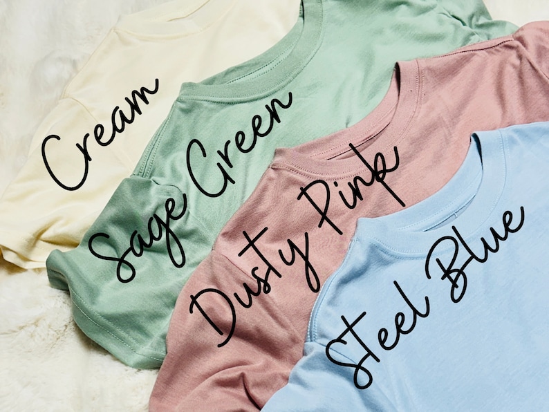 May include: A collection of five crew neck t-shirts in various pastel colors: cream, sage green, dusty pink, and steel blue. The names of the colors are written in cursive across the shirts.