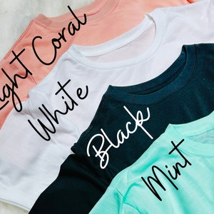May include: A close-up shot of four t-shirts in different colors: light coral, white, black, and mint. The names of the colors are written in cursive on each shirt. The shirts are neatly folded, showcasing their crew necklines and short sleeves.