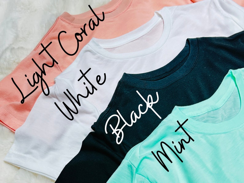 May include: A close-up shot of four t-shirts in different colors: light coral, white, black, and mint. The names of the colors are written in cursive on each shirt. The shirts are neatly folded, showcasing their crew necklines and short sleeves.