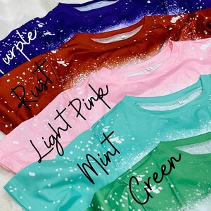 May include: An assortment of crew neck t-shirts in shades of purple, rust, light pink, mint, and green. Each top features a bleached, speckled pattern, with the colour name written across the front.