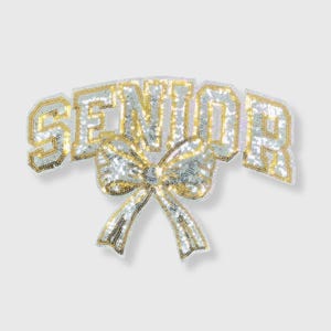 May include: A sequined patch with the word "SENIOR" in white and gold lettering, arched above a large bow. The bow and letters are outlined in gold, creating a celebratory design.